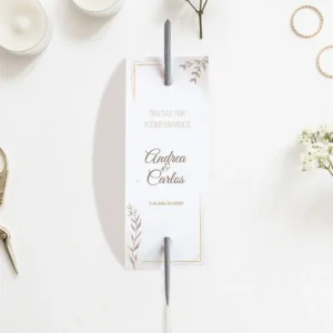 Etiqueta para bengalas de boda con diseño minimalista en blanco y detalles dorados, personalizada con los nombres Andrea & Carlos y la fecha del evento, colocada sobre una mesa decorada con velas y flores.