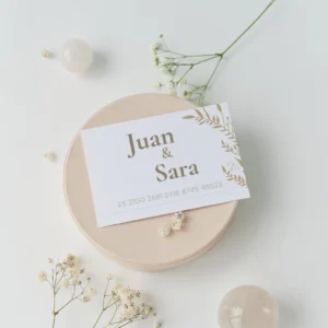 Tarjeta de regalo con nombres 'Juan & Sara', diseño floral dorado sobre fondo blanco, colocada sobre una caja beige con decoración de flores secas y piedras en tonos neutros.