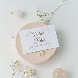 Tarjeta de cuenta bancaria para regalo de boda con los nombres 'Andrea & Carlos' y número IBAN, decorada con un marco dorado en estilo clásico y elegante, sobre una base clara con flores blancas alrededor.