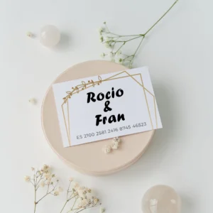 Tarjeta de cuenta bancaria para regalo de boda con los nombres 'Rocío & Fran' y número IBAN, decorada con un marco geométrico dorado y detalles de hojas, sobre una base clara con flores blancas alrededor.