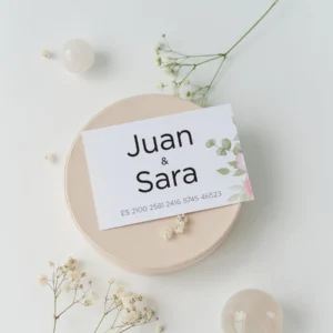 Tarjeta de cuenta bancaria para regalo de boda con los nombres 'Juan & Sara' y número IBAN, decorada con un diseño floral en tonos verdes y rosados, colocada sobre una base clara con flores blancas alrededor.