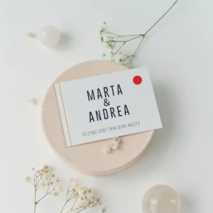 Tarjeta de cuenta bancaria para regalo de boda con los nombres 'Marta & Andrea' y número IBAN, diseñada en estilo minimalista con detalles dorados y un círculo rojo, decorada con flores blancas sobre fondo claro.