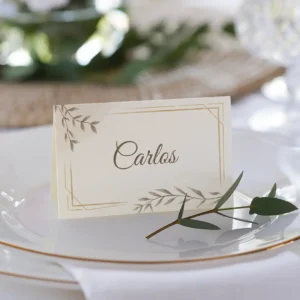 Tarjeta de lugar para boda con el nombre 'Carlos', diseño en tonos beige con detalles dorados y motivos de hojas, colocada sobre un plato blanco con borde dorado y decorada con una ramita de olivo.