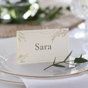 Tarjeta de sitio personalizada con el nombre 'Sara', diseñada en tonos beige y dorado con detalles florales, colocada sobre un plato blanco con borde dorado en una mesa decorada para boda.