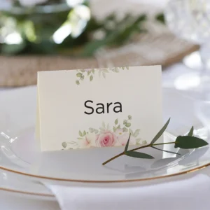 Tarjeta de lugar para boda con el nombre 'Sara', diseño floral en tonos verdes y rosas, colocada sobre un plato blanco con borde dorado y decorada con una ramita de olivo.