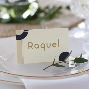 Tarjeta de lugar para boda con el nombre 'Raquel', diseño en tonos beige, dorado y azul marino, colocada sobre un plato blanco con borde dorado y decorada con una ramita de olivo.