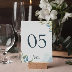Número de mesa '05' para boda con diseño floral en tonos azules y verdes, acompañado de los nombres 'Marta & Oliver'. Colocado sobre un soporte de madera en una mesa decorada con cristalería, flores blancas y velas.