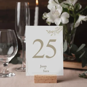 Número de mesa '25' para boda con diseño elegante en tonos dorados y detalles de hojas, acompañado de los nombres 'Juan & Sara'. Colocado sobre un soporte de madera en una mesa decorada con cristalería, flores blancas y velas.