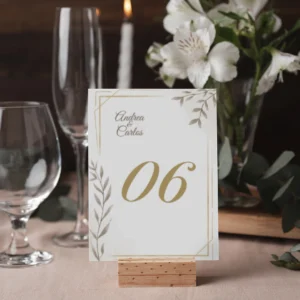 Mesero de boda con diseño elegante en tonos beige y dorado, número '06' en tipografía sofisticada, personalizado con los nombres Andrea & Carlos, colocado sobre un soporte de madera en una mesa decorada con flores blancas y cristalería.