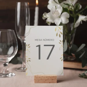 Mesero de boda con diseño floral minimalista, fondo blanco y detalles dorados, sobre una mesa decorada con copas, flores blancas y velas.