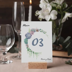 Mesero de boda con diseño floral de suculentas y detalles verdes, número '03' en tipografía elegante, personalizado con los nombres de los novios, colocado sobre un soporte de madera en una mesa decorada con flores y cristalería.