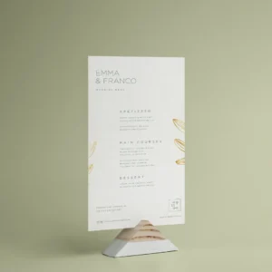 Menú de boda personalizado con los nombres 'Emma & Franco', diseñado en estilo minimalista con detalles dorados sobre fondo blanco. Presentado en un soporte geométrico sobre fondo verde suave.