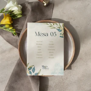 Tarjeta de asignación de mesa para boda con diseño floral en tonos azules y verdes, con nombres de los invitados y detalles elegantes, colocada sobre un plato de cerámica con decoración en tonos neutros y flores frescas.