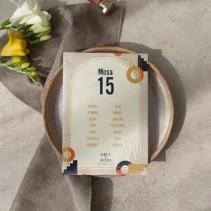 Tarjeta de asignación de mesa para boda con diseño art déco en tonos beige, azul marino y dorado, con nombres de los invitados, colocada sobre un plato de cerámica con decoración en tonos neutros y flores frescas.