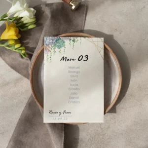 Tarjeta de asignación de mesa para boda con nombres de los invitados, diseño floral con suculentas y detalles dorados, colocada sobre un plato de cerámica con decoración en tonos neutros y flores frescas