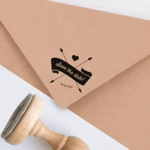 Sello de caucho con la frase 'Save the Date' en una cinta decorativa, acompañado de flechas cruzadas y un corazón, estampado en tinta negra sobre un sobre kraft, junto a un sello de madera