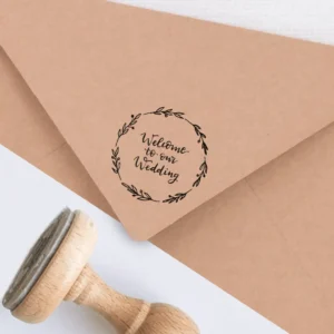 Sello de caucho con la frase 'Welcome to our Wedding' rodeada de una corona de hojas, estampado en tinta negra sobre un sobre kraft, acompañado de un sello de madera.