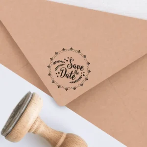 Sello de caucho con la frase 'Save the Date' rodeada de una corona floral, estampado en tinta negra sobre un sobre kraft, junto a un sello de madera.