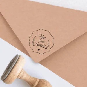 Sello de caucho con la frase 'You are Invited' en un diseño vintage con detalles de espigas y un pequeño corazón, estampado en tinta negra sobre un sobre kraft, junto a un sello de madera.